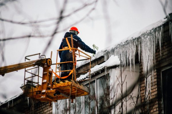 A worker in a red helmet stands on a lift, wearing cold weather PPE, as he clears snow and ice from the edge of a building roof. Large icicles hang perilously, while the scene is framed by blurred tree branches, capturing the essence of a frigid winter day.