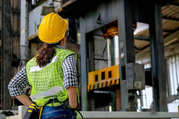 A person wearing a yellow hard hat and a fluorescent green safety vest stands with their back to the camera, observing heavy machinery in an industrial setting. The individual appears to be monitoring the point of operation, ensuring the equipment functions safely and efficiently.