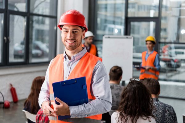 A man wearing an orange safety vest and red hard hat holds a blue clipboard and smiles at the camera. Behind him, a group of people are attending a workplace safety training session, with another person in an orange vest and yellow hard hat presenting near a whiteboard, ensuring OSHA standards are met.