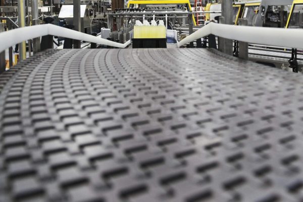 A close-up view of conveyor belts in an industrial factory setting, with machinery and production equipment visible in the background. The conveyor swiftly transports a small object, while the surrounding area is illuminated with industrial lighting.