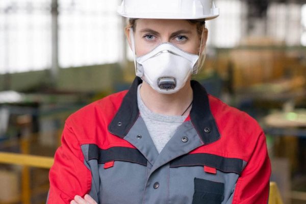 A person wearing a white hard hat and a gray and red work uniform stands confidently with arms crossed in an industrial setting. As the working lead, they're also donning a protective face mask amidst the background of machinery and equipment.