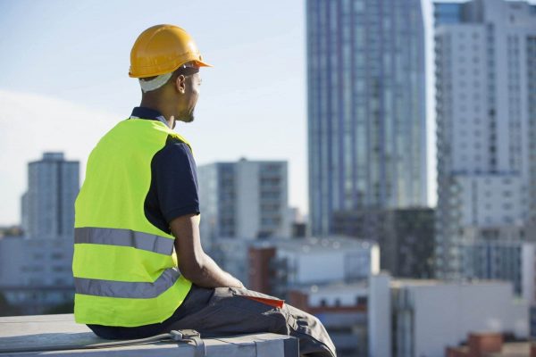 A construction worker wearing a yellow safety vest and a hard hat sits on the edge of a building, gazing towards a cityscape with high-rise buildings in the background. Amidst the clear daytime sky, one can't help but wonder about the stress on the job that accompanies such heights.