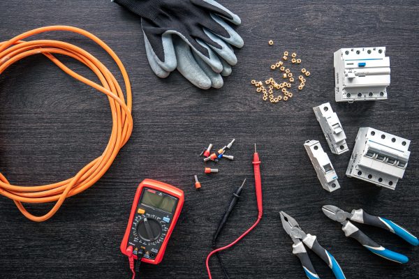 A variety of electrical tools and components are spread out on a dark surface. There is an orange cable, a pair of gloves, essential electrical safety equipment like a digital multimeter, wire cutters, and several circuit breakers. Additionally, there are small screws and various electrical connectors.