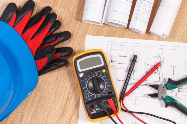 A multimeter, wires, and a pair of pliers rest atop construction blueprints. Nearby, a blue hard hat and red-and-black work gloves are positioned, highlighting the importance of electrical safety. The scene suggests an ongoing electrical or construction project with rolled-up blueprints also visible.