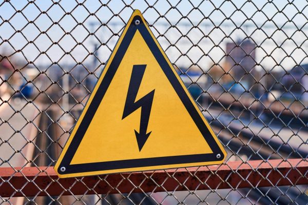 A triangular yellow warning sign with a black lightning bolt symbol is attached to a chain-link fence, indicating a danger of electrical hazards. The background is blurred, showing structures and an industrial environment, reminding everyone to stay safe around electricity.