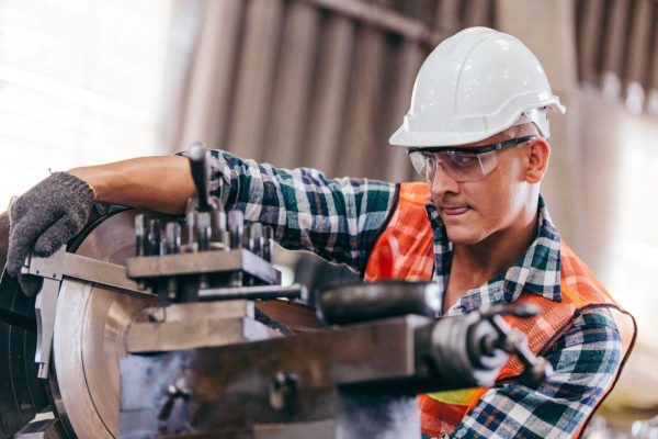 A man wearing a hard hat, safety glasses, and a checkered shirt with a safety vest is operating a large industrial machine. Focused on lathe safety, he uses his gloved hands to handle the equipment meticulously in a well-lit workshop.