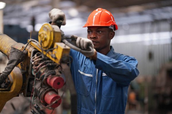 An individual wearing a blue coverall and an orange hard hat is operating or repairing industrial machinery. They are focused on their task, using both hands to work on the equipment in what appears to be a factory or workshop environment, while carefully ensuring the machine guard is properly in place.