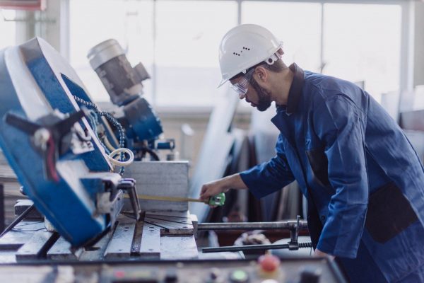 A person wearing a white hard hat, safety glasses, and a blue work uniform operates machinery in an industrial workshop. They are adjusting equipment and working with various tools on a metal surface, with background equipment and tools visible in the workshop.