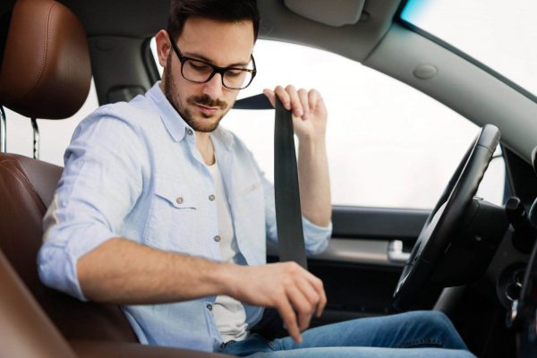 A man with glasses and a beard, wearing a light blue shirt and jeans, sits in the driver's seat of a modern car with brown leather seats. With his right hand, he fastens his seatbelt, emphasizing the importance of safety belts as he looks down to secure it properly.