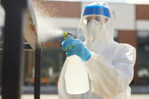 A person dressed in full protective gear, including a white hazmat suit, high-visibility clothing, blue gloves, face mask, and face shield, is spraying disinfectant from a spray bottle onto surfaces. The background shows an outdoor setting with a blurry building.