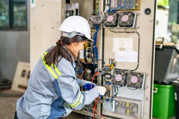 A person wearing a white hard hat and a gray jacket with reflective stripes is diligently working on an open electrical panel, engaged in industrial electrical installation. They are using a handheld tool with wires connected to the panel. Cooling fans, cables, and various components are visible inside the panel.