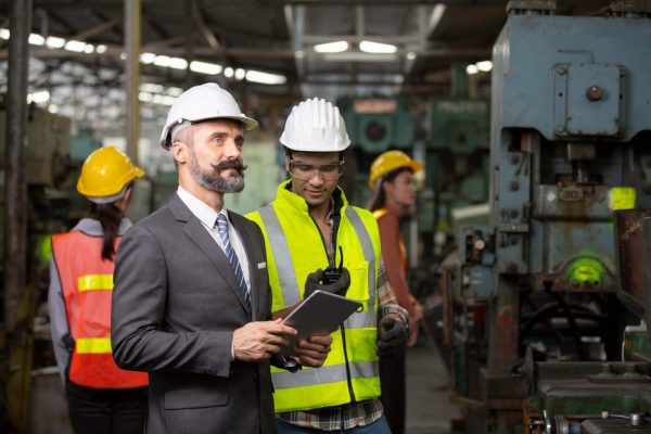 A bearded man in a suit and hard hat holds a tablet while standing in a factory, discussing employee compliance with a worker in a high-visibility vest and hard hat. Two other workers in high-vis vests and hard hats are seen in the background amid industrial machinery, ensuring safety culture is maintained.