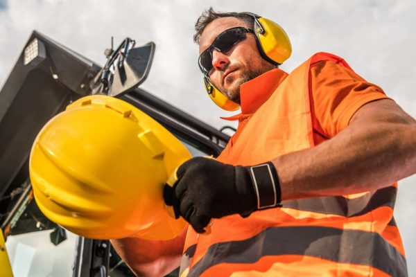 A construction worker wearing an orange safety vest, black gloves, and yellow ear protection holds a yellow safety helmet, emphasizing head protection. The worker also has sunglasses on, and heavy machinery is visible in the background under a cloudy sky.