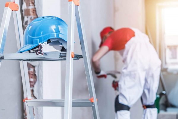 A blue hard hat rests on top of an aluminum ladder in the foreground, emphasizing ladder safety at the workplace. In the background, a construction worker in a white coverall and red shirt works on a wall, slightly out of focus. Sunlight filters through a nearby window.