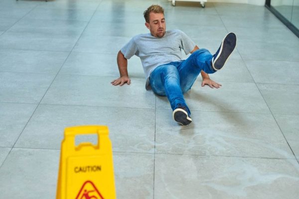 A man slips and falls on a wet floor in a hallway. He appears to be losing his balance while sitting on the floor, with one leg in the air. A yellow caution sign indicating "Wet Floor" is prominently placed in the foreground.