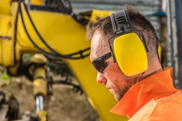 A construction worker is wearing yellow earmuffs for hearing protection, safety sunglasses, and an orange high-visibility jacket. The worker is standing close to a piece of heavy yellow machinery, which is blurred in the background.