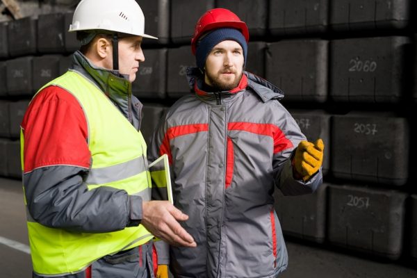 Two construction workers in safety gear stand in front of stacked blocks. One, wearing a white helmet and yellow vest, holds a tablet, emphasizing essential training. The other, in a red helmet and gray jacket, gestures with one hand while discussing how to stay safe on the site.