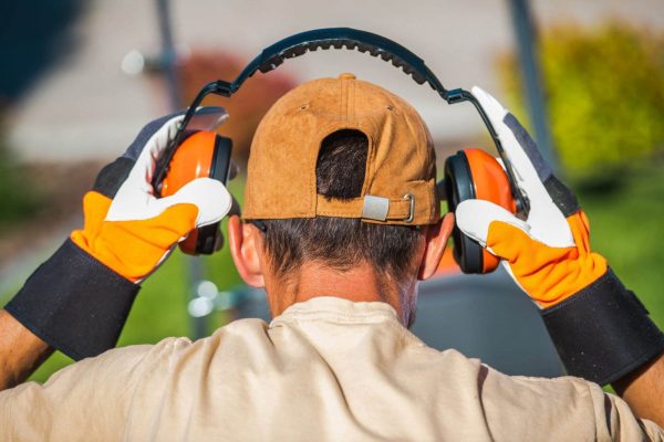 A man wearing a brown cap, beige shirt, and orange work gloves adjusts his orange and black hearing protection earmuffs, viewed from behind. The background is out of focus, suggesting an outdoor setting with green foliage.