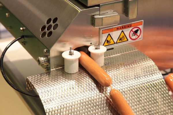 A machine in operation with hot dogs conveyed through it. The metal surface is perforated, and two white rollers are positioned above the hot dogs. Warning labels are visible, indicating caution and safety instructions to prevent any risk of injury.