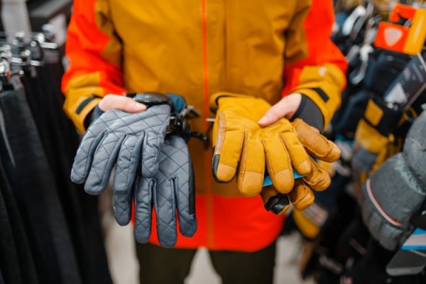 A person in an orange jacket holds two pairs of gloves, perfect for cold weather jobs: a quilted blue pair in the left hand and a tan pair in the right. The store's background displays various options to help you layer up against the cold.