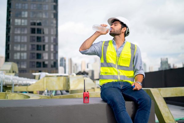 A construction worker wearing a white helmet, yellow safety vest, blue shirt, and jeans sits on a concrete ledge. He is drinking water from a bottle, with a walkie-talkie placed beside him. Emphasizing OSHA Heat Standards in hot environments, the cityscape with high-rise buildings is visible in the background.