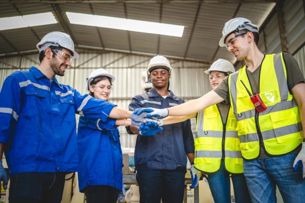 A group of five industrial workers, wearing hard hats and safety gear, stand in a circle inside a factory or warehouse. They are smiling and stacking their hands together in the center, showcasing teamwork and unity during a PPE training session.