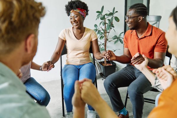 A group of diverse individuals sit in a circle holding hands and smiling. They are in a brightly lit room with a potted plant in the background. The atmosphere is positive and supportive, suggesting a workplace mental health activity or support meeting.