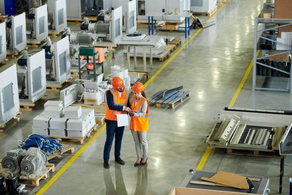 Two workers wearing orange safety vests and hard hats are standing in a spacious industrial warehouse, discussing a document. The warehouse floor is lined with various machinery and equipment, with aisles giving access to different sections of the facility.