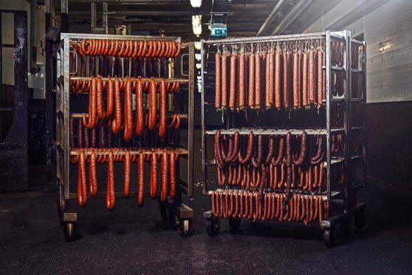 Two metal racks filled with various types of hanging sausages in a dimly lit industrial setting look like the handiwork of a skilled slaughterhouse employee. The sausages hang in neat rows, showcasing different shapes and sizes, illuminated by overhead lights. The background features industrial pipes and walls.