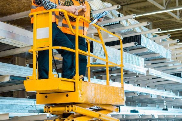 Two construction workers wearing orange helmets and safety vests are standing on a yellow scissor lift, examining documents. They are in a large warehouse with metal beams and building materials on shelves in the background, meticulously planning their next task.