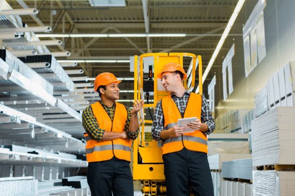 Two men wearing orange safety vests and hard hats are standing in a warehouse aisle. One man is holding a clipboard and the other is holding a walkie-talkie. They are smiling and talking to each other, discussing OSHA guidelines, surrounded by shelves filled with materials.