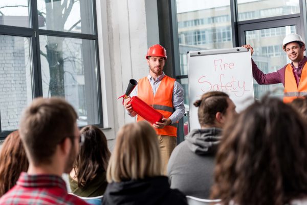 Two men wearing safety vests and helmets are conducting a comprehensive guide to fire safety training. One is holding a fire extinguisher, while the other stands near a whiteboard with "Fire Safety" written on it. They are speaking to a seated audience in a bright room.