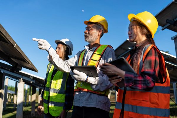 Three engineers wearing safety helmets and reflective vests are inspecting solar panels at a solar farm, showcasing excellent workplace safety. The person in the middle is pointing towards something while the other two are holding tablets. The sky is clear and blue.