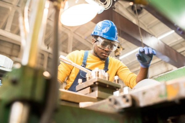 A worker wearing a blue hard hat, safety goggles, blue gloves, and a yellow shirt operates machinery in an industrial setting. The worker appears focused on the task while adhering to lockout procedures. Various metal components and tools are visible around the workstation. Overhead, a light illuminates the area.