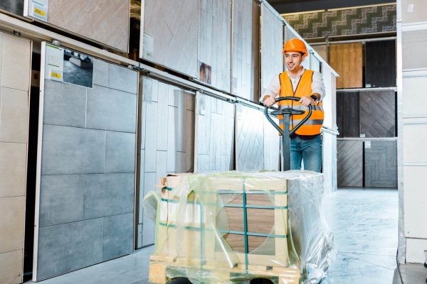 A worker in an orange safety vest and helmet navigates a pallet jack loaded with wrapped materials down a hardware store aisle lined with various types of tile samples.