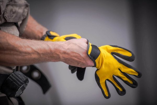 Close-up of a person wearing bright yellow safety gloves with black accents, adjusting the strap of one glove with the other hand. The person is dressed in a grey uniform with a visible utility belt. The background is blurred and neutral in color.