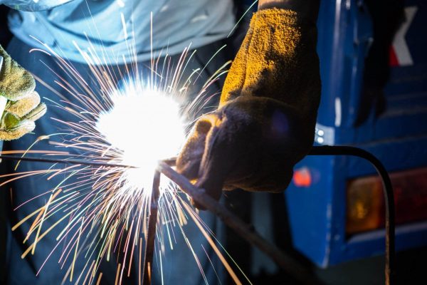 Close-up of a person welding and cutting metal at the work site, with bright sparks flying from the welding torch. The individual is wearing protective gloves and a blue shirt, working in a dimly lit environment. The background shows a blurred blue object.