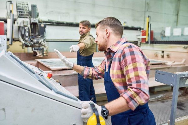 Two factory workers operate machinery in an industrial setting, focused on accidents prevention. One worker, in a plaid shirt and safety gloves, presses buttons on a control panel, while the other, in a brown shirt and blue overalls, gestures with his hand in the background.