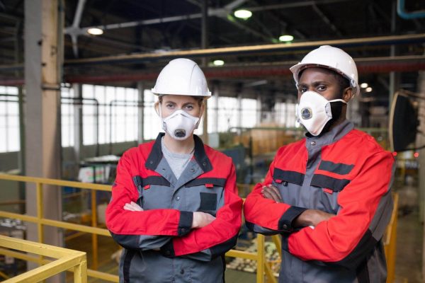 Two factory workers, wearing white hard hats and protective face masks, stand with arms crossed in an industrial setting. Dressed in grey and red work uniforms, they emphasize the importance of making respiratory protection a priority while surrounded by machinery in a large warehouse.